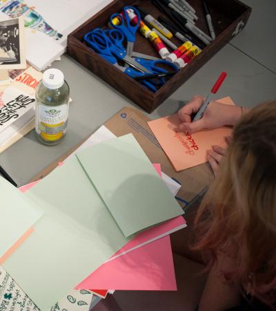 person writing on booklet on table covered in supplies