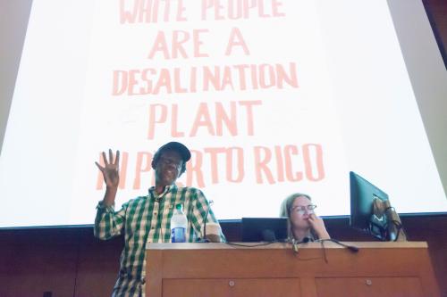 artist Pope L at lectern in front of projection screen